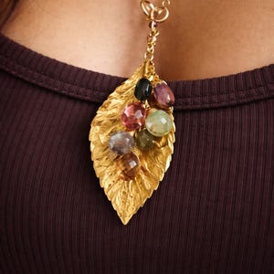 Puede incluir: Un collar con colgante de hoja dorada con un grupo de gemas coloridas. Las gemas incluyen piedras rosas, verdes, moradas y marrones. El collar se lleva sobre una camiseta marrón oscuro.