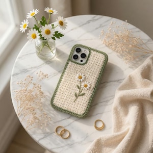 May include: A cream crochet phone case with a daisy design. The case has a green border and embroidered daisies. The phone case is on a marble table with a small vase of daisies and gold rings.