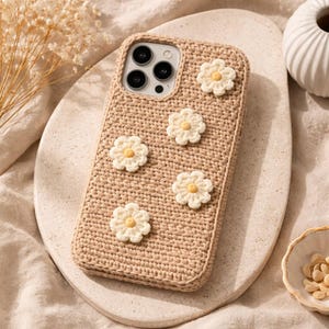 May include: A beige crochet phone case decorated with five white daisy appliqués with yellow centres. The case has a woven texture and is designed to fit a smartphone. The daisies are evenly spaced across the surface.