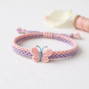 Puede incluir: Pulsera hecha a mano con diseño de punto rosa y lavanda. La pulsera tiene un adorno de mariposa en el centro, con cuerpo azul claro y alas rosas. Una delicada pieza de joyería.