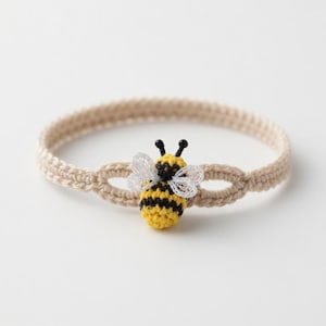 May include: A beige crochet headband with a yellow and black bee applique. The bee has white wings and black antennae. The headband is made of a soft, woven material, suitable for all ages.