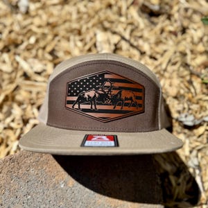 May include: A tan and brown baseball cap featuring a hexagonal patch on the front panel. The patch displays a black graphic of two elk locking antlers against a background of the American flag. A small rectangular sticker is on the flat brim.