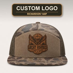 May include: A camouflage trucker hat with a brown leather patch featuring an owl graphic and the text "GREAT HORNED PRECISION". The hat has a flat brim and mesh back panels. Above the hat, a rectangular sign reads "CUSTOM LOGO" and "RICHARDSON 168P".