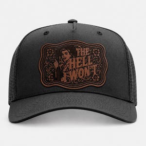 May include: A charcoal gray trucker hat featuring a brown leather-like patch on the front. The patch displays an engraved illustration of a person holding a firearm next to a cactus, surrounded by floral patterns and the text THE HELL I WON'T.