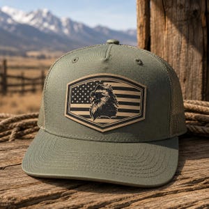 May include: An olive green trucker hat featuring a structured front panel and a mesh back. A tan leather-like patch is centered on the front, displaying a laser-engraved graphic of an eagle wearing sunglasses superimposed over an American flag.