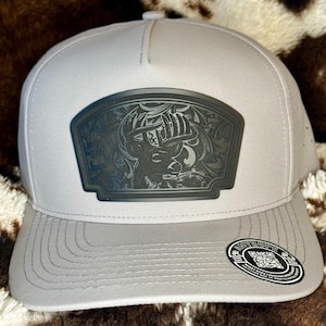 May include: A light gray trucker hat with a dark gray patch featuring a detailed buck deer design. The hat has a curved brim and a mesh back. The patch has an ornate border and the hat has a snap closure.