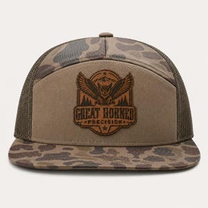 May include: A camouflage print trucker hat with a solid tan front panel featuring a brown leather patch. The patch displays an engraved graphic of an owl with spread wings above a forest and mountains, with the text GREAT HORNED PRECISION.
