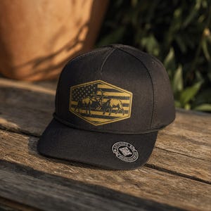 May include: Black baseball cap with a gold and black emblem featuring two bull elk and the American flag. The cap has a curved brim and a black rope detail. The cap is sitting on a wooden surface.