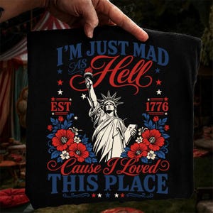 May include: A black t-shirt featuring a graphic of the Statue of Liberty holding a torch. The text reads I'M JUST MAD AS HELL EST 1776 CAUSE I LOVED THIS PLACE. The design includes red and blue floral accents and stars on a dark background.