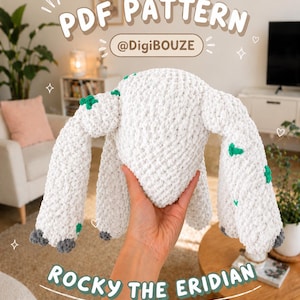 Puede incluir: Una criatura blanca y afelpada, tejida a ganchillo, con detalles verdes y grises, sostenida en una mano. El texto "PDF PATTERN @DigiBouze" y "ROCKY THE ERIDIAN" son visibles. La criatura tiene brazos largos y un cuerpo redondeado.