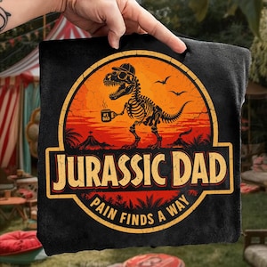 Pode incluir: Toalha quadrada preta com um gráfico Jurassic Dad. O design apresenta um esqueleto de dinossauro usando um chapéu e segurando uma caneca de café, contra um pôr do sol laranja. O texto inclui "Jurassic Dad" e "Pain Finds a Way".