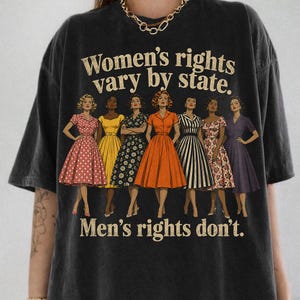 May include: A charcoal grey t-shirt featuring a graphic of seven individuals in vintage-style dresses. The text reads: Women's rights vary by state. Men's rights don't. The figures are depicted in various solid and patterned dresses including polka dots and stripes.