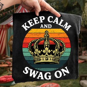 Könnte beinhalten: Schwarzes quadratisches Kissen mit dem Text "KEEP CALM AND SWAG ON" in Weiß. Das Design beinhaltet eine goldene Krone über einem Retro-Sonnenuntergangshintergrund aus orangefarbenen, gelben und türkisfarbenen Streifen. Das Kissen wird von einer Hand gehalten.