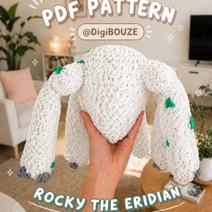 Puede incluir: Un juguete de peluche de ganchillo blanco y texturizado llamado "Rocky the Eridian" con detalles verdes y grises. La imagen incluye el texto "PDF PATTERN" y el nombre de usuario del vendedor, "@DigiBouze". El juguete es sostenido por una mano.
