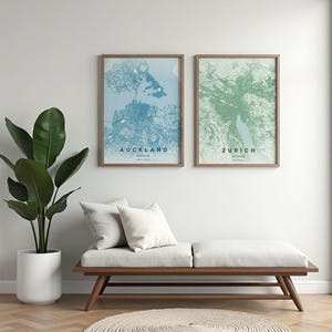 May include: Two framed city map prints, one blue and one green, hang above a wooden bench with white cushions. The blue map is of Auckland, New Zealand, and the green map is of Zurich, Switzerland. A large potted plant is to the left.