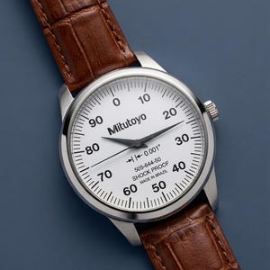 May include: A silver-cased Mitutoyo watch with a white face and black numerals. The watch has a brown leather band. The face displays the text "Mitutoyo", "0.001" and "SHOCK PROOF MADE IN BRAZIL".