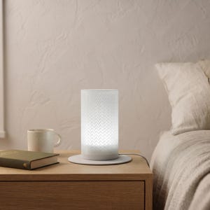 May include: A cylindrical table lamp with a white, translucent shade featuring a repeating chevron pattern. The lamp emits a soft, diffused light and sits on a circular base with a power cord extending from the side, resting on a wooden surface.