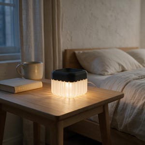 May include: A square-shaped electric lamp with a black top and a translucent, vertically ribbed base that emits a warm, soft glow. The lamp is positioned on a wooden surface next to a closed book and a ceramic mug, casting light downward.