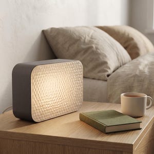 May include: A rectangular table lamp with a dark gray outer shell and a textured, translucent front panel that emits a warm, soft glow. The lamp features rounded corners and a smooth finish, providing illumination through its patterned surface.