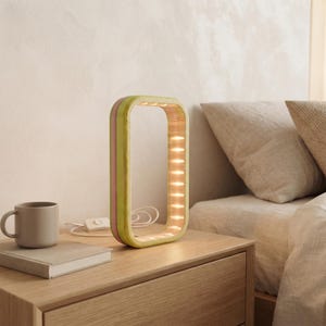 May include: A rectangular table lamp with a rounded frame made of layered wood in shades of light green and pink. The inner edge of the frame features a series of small, warm-toned LED lights. A white power cord with a switch extends from the base.