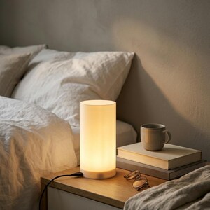 May include: A cylindrical table lamp with a white frosted glass shade emitting a warm glow, connected by a black power cord. The lamp sits on a wooden surface next to a stack of two books, a ceramic mug, and a pair of metal-framed eyeglasses.