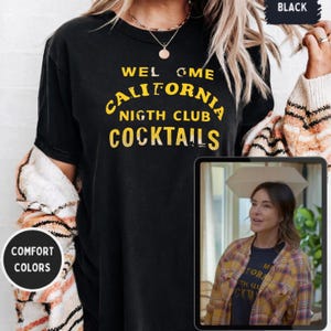 May include: Black t-shirt with the text "WELCOME CALIFORNIA NIGHT CLUB COCKTAILS" in yellow lettering. The shirt has a crew neck and short sleeves. The words "COMFORT COLORS" are in a white circle.