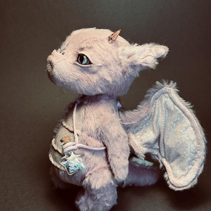 Puede incluir: Un dragón de peluche en un suave tono lavanda, con alas texturizadas de acabado plateado brillante. La criatura lleva un pequeño colgante transparente en forma de estrella relleno de purpurina azul sujeto por una correa y tiene un cuerno.