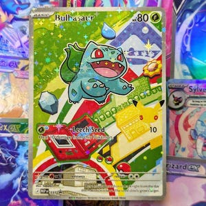 Könnte beinhalten: Eine holografische Bulbasaur Pokémon-Sammelkarte. Die Karte hat einen grünen Hintergrund mit einem Cartoon-Bulbasaur-Charakter. Die Karte trägt den Text "Bulbasaur" und "Leech Seed". Weitere Karten sind im Hintergrund sichtbar.