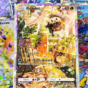 May include: A holographic Rowlet Pokémon trading card. The card features a colorful illustration of Rowlet in a hanging basket. The card has the text "Rowlet" and "Fury Attack" with damage details. Other cards are visible in the background.