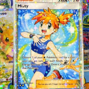 May include: A holographic "Misty" Trainer card from the Pokémon trading card game. The card features a smiling anime character with orange hair and a blue outfit, set against a sparkling blue background. The card is encased in a protective sleeve.