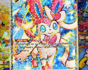 May include: A holographic Sylveon EX Pokémon trading card. The card features a pink and blue Sylveon character with the text "Sylveon EX" and "Happy Ribbon". The card has a HP of 140 and a Fairy type attack.