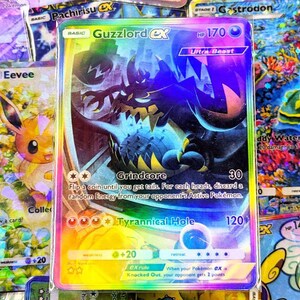 May include: A holographic Guzzlord ex Pokémon trading card. The card features a dark purple and black monster with the text "Guzzlord ex" at the top. The card has a rainbow holographic background and is surrounded by other Pokémon cards.