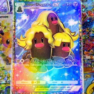 May include: A holographic Alolan Dugtrio EX Pokémon card. The card features three Dugtrio characters with yellow hair and pink noses against a blue starry background. The card has text that reads "Triplet Headbutt" and "HP 130".