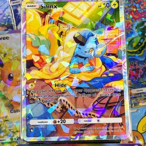Könnte beinhalten: Eine holografische Shinx Pokémon-Sammelkarte. Die Karte zeigt einen blauen Shinx-Charakter mit gelben Akzenten vor einem farbenfrohen Hintergrund. Die Karte enthält Text, einschließlich des Namens "Shinx" und Spielanweisungen.