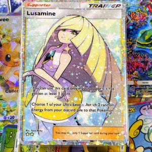 May include: A holographic Pokemon trading card featuring Lusamine, a Trainer card. The card has a silver border and a colorful illustration of a woman with long blonde hair. Text on the card includes "Supporter" and game instructions.