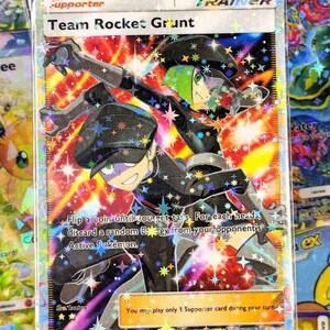 Może przedstawiać: Holograficzna karta Trenera Team Rocket Grunt. Karta przedstawia dwie postacie w czarnych mundurach z zielonymi i fioletowymi akcentami, na tle kolorowego, usianego gwiazdami tła. Tekst zawiera "Team Rocket Grunt" i instrukcje gry.