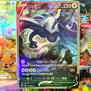Può includere: Carta collezionabile Pokémon olografica con Lugia V. La carta ha un bordo argentato e mostra un'illustrazione dettagliata di Lugia in volo, con il testo "Read the Wind" e "Aero Dive". La carta ha 220 HP.