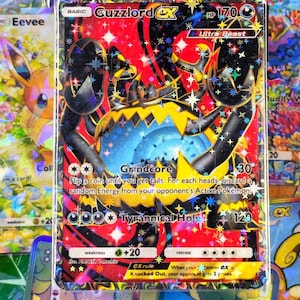 May include: A Guzzlord EX Pokémon trading card with a red, black, and yellow design. The card features the text "Guzzlord EX" and "Ultra Beast". The card has a holographic, sparkly finish and is surrounded by other Pokémon cards.