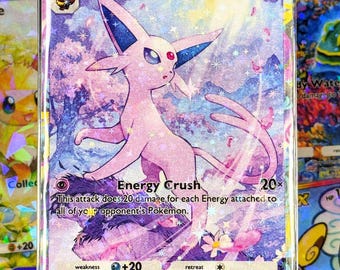 May include: A holographic Espeon Pokémon card. The card features a pink Espeon character with blue accents, set against a background of purple and pink hues. The card includes text such as "Espeon" and "Energy Crush".