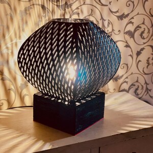 Pode incluir: Uma lâmpada de mesa preta e geométrica com uma sombra com padrão de diamante. A lâmpada está sobre uma base retangular preta com um detalhe vermelho. A lâmpada está sobre uma mesa branca, com sombras.