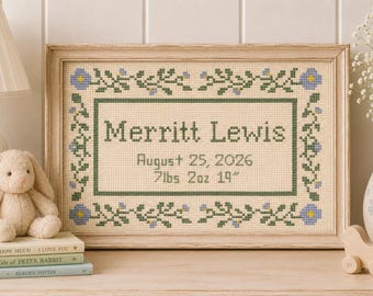 May include: A framed cross-stitch birth announcement featuring the name Merritt Lewis, the date August 25, 2026, and the birth stats 7lbs 2oz 19 inches. The design includes a border of green vines with small blue flowers on a cream fabric background.