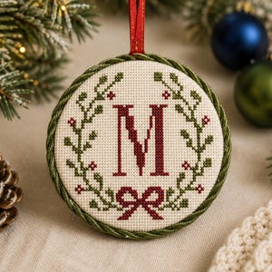 Puede incluir: Un adorno navideño redondo con un diseño de punto de cruz sobre tela color crema. El centro muestra una letra M roja grande rodeada por una corona de hojas verdes con pequeñas bayas rojas y un lazo rojo en la parte inferior. Borde de cordón verde.