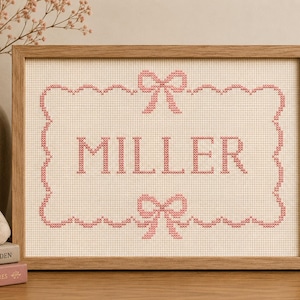 Può includere: Una cornice rettangolare in legno contenente un motivo a punto croce su tessuto color crema. Il design presenta il nome MILLER in lettere maiuscole rosa, racchiuso all'interno di un bordo smerlato decorato con un fiocco rosa al centro.
