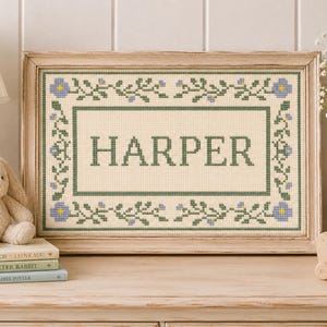 Op de afbeelding: Een rechthoekig kruissteek borduurwerk ingelijst in licht hout, met de naam HARPER in donkergroene blokletters gecentreerd binnen een decoratieve rand van groene ranken, bladeren en kleine blauwe bloemen op een crèmekleurige stof.