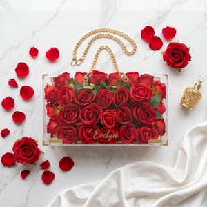 May include: A clear acrylic handbag filled with red roses, featuring a gold chain handle. Scattered rose petals and a gold perfume bottle are arranged on a marble surface. The name "Evelyn" is inscribed on the bag.