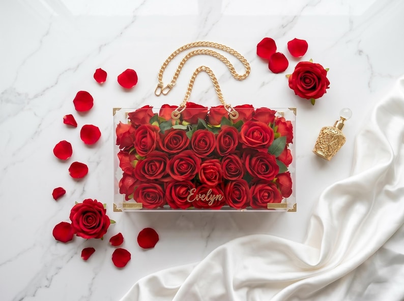 May include: A clear acrylic handbag filled with red roses, featuring a gold chain handle. Scattered rose petals and a gold perfume bottle are arranged on a marble surface. The name "Evelyn" is inscribed on the bag.