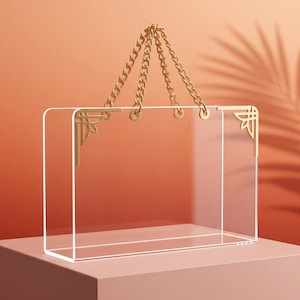 May include: A rectangular clear acrylic handbag featuring gold-toned metal chain handles and decorative gold-toned corner accents. The bag has a transparent body with a structured design, connected to the chain handles by circular gold-toned hardware.