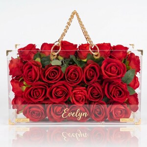May include: A clear acrylic handbag filled with vibrant red roses. The bag has a gold chain handle and gold corner accents. The name "Evelyn" is displayed on the front. The bag is filled with red roses and green leaves.