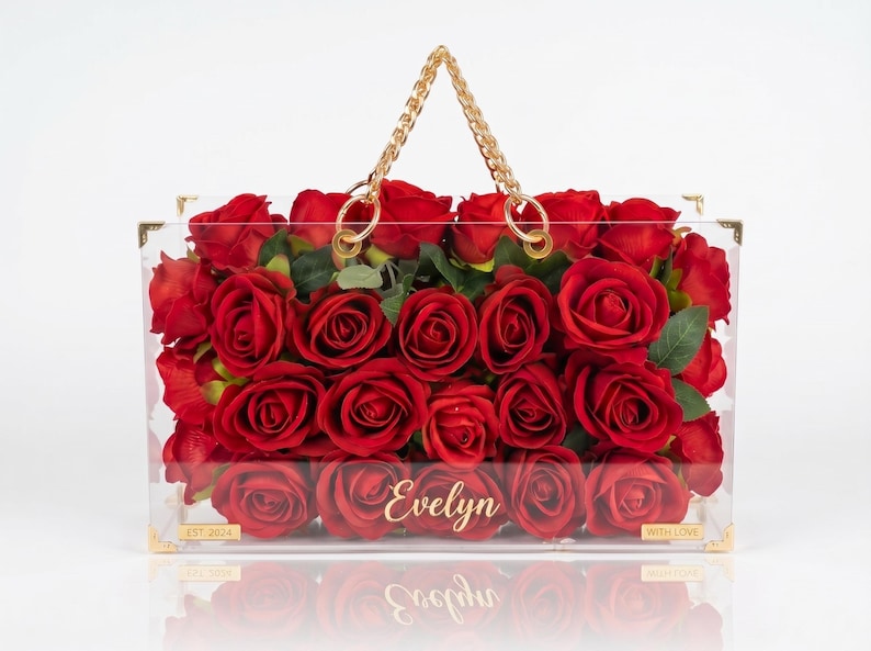May include: A clear acrylic handbag filled with vibrant red roses. The bag has a gold chain handle and gold corner accents. The name "Evelyn" is displayed on the front. The bag is filled with red roses and green leaves.