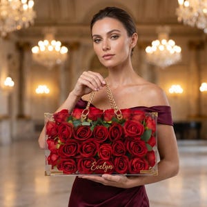 May include: A clear rectangular handbag with gold trim and a gold chain handle, filled with vibrant red roses. The name "Evelyn" is inscribed on the front. The bag is held against a blurred background of a grand hall.